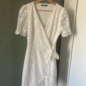 Vit omlottklänning från Gina Tricot  - Superfin vit omlottklänning från Gina tricot med korta puffärmar och vackert broderat blommönster. Klänningen har knyt i midjan och v-ringning, perfekt för sommarens alla tillfällen. Lätt och luftig känsla!