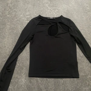 Svart långärmad topp med cut out BikBok - Svart långärmad topp från BikBok med en snygg cut out-detalj vid halsen och med glansigt skönt tyg. Modellen är figurnära och har en enkel, stilren look som passar perfekt till jeans eller kjol. Mjuk och stretchig känsla, perfekt för dig som gillar trendiga detaljer.