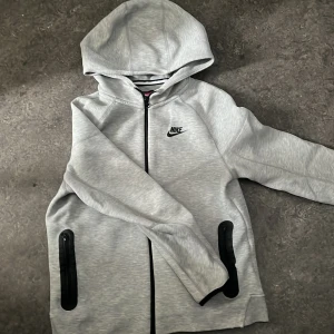 Grå tracksuit från Nike - Snygga grå mjukisbyxor från Nike med svart snörning i midjan och elastiska muddar vid bensluten. Byxorna har en sportig look med diskret Nike-logga på ena benet och är tillverkade i mjukt bomullsmaterial. Perfekta för chill eller träning.