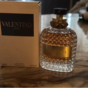 Valentino Uomo 100ml - Lyxig herrparfym från Valentino, Uomo Eau de Toilette i 100 ml flaska. Flaskan är mönstrad i glas med guldiga detaljer och svart sprayhuvud. Doften är elegant och flaskan har en exklusiv känsla med sin guldplatta och geometriska design.