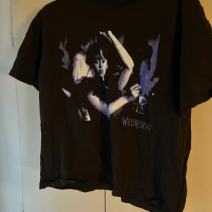 Svart Wednesday t-shirt från Divided - Cool svart t-shirt från Divided med tryck av Wednesday Addams i lila och vitt på framsidan. Klassisk passform med rund hals och korta ärmar. Perfekt för dig som gillar statement-plagg och popkultur.