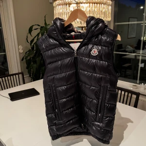 Moncler Mörkblå dunväst med luva - Snygg mörkblå dunväst från Moncler med glansig finish, vadderad och med dubbla luvor. Använd 1 gång och är helt felfri i nyskick. Köpt på NK i januari kvitto finns.