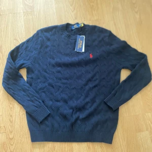 Mörkblå stickad tröja Polo Ralph Lauren - Mörkblå stickad tröja från Polo Ralph Lauren med diskret strukturmönster och klassisk röd logga broderad på bröstet. Tröjan har rund halsringning, långa ärmar och ribbade muddar. Perfekt för dig som gillar stilren och tidlös design.
