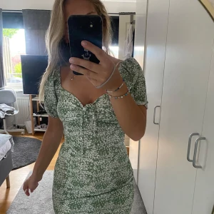 Grön blommig kortklänning Gina Tricot - Supersöt grön kortklänning från Gina Tricot med vita blommor, puffärmar och söt snörning framtill. Klänningen har figurnära passform och är perfekt för sommarens alla tillfällen. Materialet är mjukt och lätt, vilket gör den riktigt skön att bära. Använd 1 gång