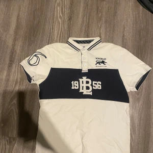 Vit och marinblå piké från Bondelid - Snygg pikétröja från Bondelid i vit och marinblå med broderade detaljer och tryck framtill. Klassisk krage med knappar och randiga detaljer på krage och ärmslut. Perfekt för en sportig och stilren look.