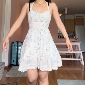 Blommig volangklänning med spetsdetaljer - Supersöt kortklänning i ljusbeige med blommigt mönster i blått och rosa. Klänningen har volanger, spetsdetaljer och knytband i ryggen. Perfekt för sommarens alla tillfällen! Materialet är mjukt och luftigt, vilket gör den extra bekväm.