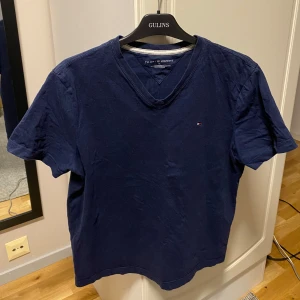 Marinblå t-shirt från Tommy Hilfiger - Snygg marinblå t-shirt från Tommy Hilfiger med liten broderad logga på bröstet. Klassisk v-ringning. Tillverkad i mjuk bomull som känns skön mot huden. Perfekt för en avslappnad och stilren look.