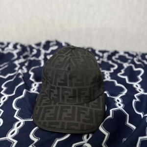 Svart keps från Fendi med FF-mönster - Snygg svart keps från Fendi med det ikoniska FF-logomönstret över hela kepsen. Klassisk form med böjd skärm och justerbar baksida. Tillverkad i ett slitstarkt tyg som känns lyxigt och trendigt. Perfekt accessoar för att lyfta din outfit. TRÄFFAS BARA I GÖTEBORG tar emot byte