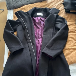 Svart kappa från Promod med dragkedja - Stilren svart kappa från Promod med snygg lila insida. Jackan har lång ärm, hög krage och stängs med dragkedja. 