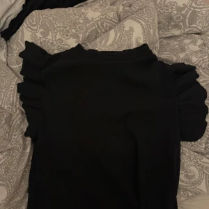 Svart topp med volangärmar - Supersnygg svart topp med volangdetaljer på ärmarna. Modellen är ribbad och har en klassisk rund halsringning. Perfekt för dig som gillar en stilren look med en twist. Passar dig som vill ha något enkelt men ändå trendigt.