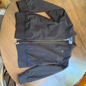 Jacka från GANT - Snygg svart bomberjacka från GANT med klassisk passform. Jackan har ribbade muddar, dragkedja framtill och två fickor. Insidan är ljusblå med stort GANT-tryck. Perfekt för dig som vill ha en stilren och tidlös look.