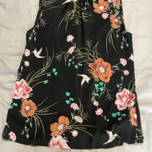 Svart blommig klänning Saint Tropez - Svart kortklänning från Saint Tropez med färgglada blommor i orange, rosa och grönt. Klänningen har ett blommigt mönster och är ärmlös, perfekt för varma dagar. Lätt och luftig modell som ger en skön känsla och snygg siluett.