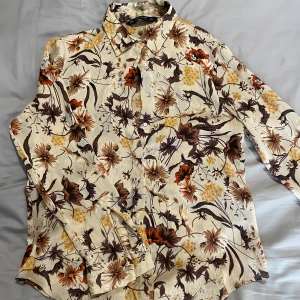 Blommig skjorta från Zara Basic M - Snygg skjorta från Zara Basic med blommigt mönster i beige, brunt, gult och orange. Skjortan har lång ärm, klassisk krage och knäppning framtill. Materialet är lätt och har en skön känsla, perfekt för dig som gillar retrovibbar och färg.