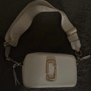 Vit axelväska Marc Jacobs - Säljer en vit axelväska från Marc Jacobs med bred axelrem där loggan är invävd. Väskan har två fack med dragkedja och snygga gulddetaljer framtill. Perfekt storlek för det viktigaste och riktigt trendig design. Den är äkta och är köpt för 2500kr. Skriv för fler bilder. PRIS KAN DISKUTERAS!!🥰