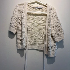 Beige stickad kofta från Zara - Superfin beige stickad kofta från Zara med kort ärm och virkade detaljer. Koftan har knytband framtill och ett unikt blommönster i strukturen. Perfekt att slänga över en topp för en chill och trendig look.