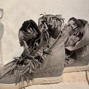 Grå mockasneakers med fransar - Unika grå sneakers i mocka med massor av fransdetaljer längs plösen och sidorna. Skorna har en platt vit sula och dekorativa sömmar runt om. Perfekta för dig som vill sticka ut med en cool och bohemisk stil.