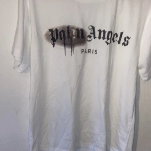 Vit Palm Angels t-shirt med tryck - Säljer en vit t-shirt från Palm Angels med ikoniskt svart logotyptryck och spray paint-effekt över bröstet. Klassisk passform och korta ärmar, perfekt för dig som gillar streetwear och vill sticka ut med en snygg statement-tee.