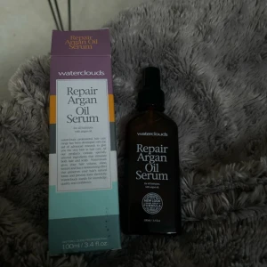 Waterclouds Repair Argan Oil Serum - Waterclouds Repair Argan Oil Serum i brun glasflaska med svart pump, 100 ml. Innehåller arganolja som ger näring och glans till håret. Kommer i originalförpackning med lila, vit och turkos design. Perfekt för att boosta håret med fukt och mjukhet.