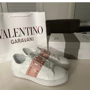 Valentino Rockstud sneakers vit/rosa - Snygga Valentino Rockstud sneakers i vitt skinn med breda ljusrosa band över vristen dekorerade med ikoniska nitar. Klassisk rund tå, platt sula och vita snören. Perfekta för dig som vill sticka ut med en lyxig och trendig look.