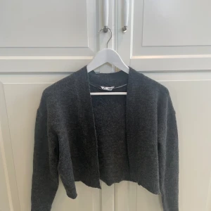 Mörkgrå stickad kofta, croppad - Mysig mörkgrå stickad kofta med croppad passform och öppen framtill. Perfekt att slänga över en t-shirt eller linne. Långärmad och enkel design som funkar till många olika stilar.