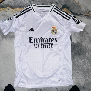 Real Madrid hemmatröja Adidas 24/25 - Snygg vit Real Madrid fotbollströja från Adidas med svarta detaljer och klubbmärke på bröstet. Tröjan har korta ärmar, svarta ränder på axlarna och Champions League-märke på ärmen. Tillverkad i lätt och ventilerande material (AEROREADY).