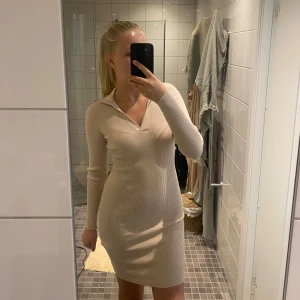 Beige ribbad kortklänning med krage - Superfin beige ribbad klänning med lång ärm och snygg krage. Klänningen är kort och har en figurnära passform som framhäver siluetten. Perfekt för dig som gillar stilrena och enkla plagg med en twist.