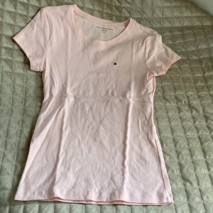 Ljusrosa t-shirt från Tommy Hilfiger - Snygg ljusrosa t-shirt från Tommy Hilfiger med klassisk rund hals och liten broderad logga på bröstet. T-shirten är i mjuk bomull och har en slim passform som sitter riktigt bra. Perfekt basic-plagg med stilren vibe.