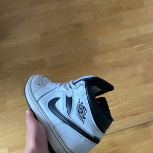 Nike Air Jordan 1 Mid svart/vit EUR 41 - Säljer ett par Nike Air Jordan 1 Mid i klassisk svart och vit färg. Skorna har svart swoosh, Air Jordan-logga på sidan och är tillverkade i skinn. De har snörning och en rund tå, perfekt för dig som gillar streetwear och sneakers med ikonisk design.