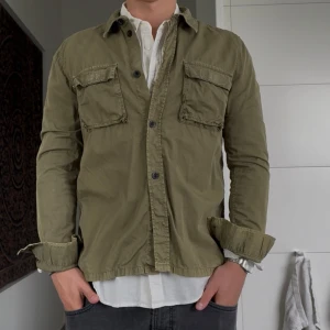 Olivgrön overshirt från Dr Denim - Olivgrön overshirt från Dr Denim med två stora bröstfickor och knappar framtill. Skjortan har lång ärm och en avslappnad passform, perfekt att bära öppen över en t-shirt eller skjorta. Tillverkad i mjuk bomull som känns skön mot huden.
