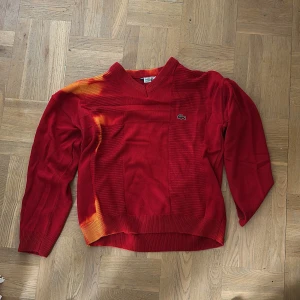Röd stickad Lacoste tröja med V-ringning,  - Röd stickad tröja från Lacoste med V-ringning och diskret krokodillogga på bröstet. Tröjan har långa ärmar och ribbade muddar. Unik orange färgskiftning på axel och nederkant ger en cool twist till looken.       Högsta bud! 