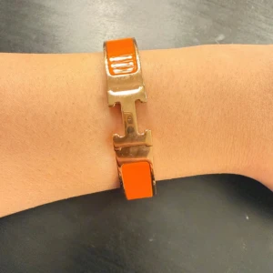 Orange armband i guld detaljer - Snyggt armband, spänne i guldfärgad metall och orange emalj. Stilren och lyxig design som verkligen sticker ut på handleden. Perfekt accessoar för dig som gillar statement-smycken och exklusiva detaljer.