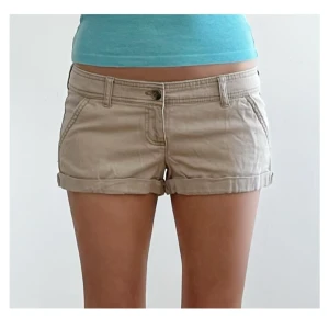 Beige shorts med upprullad kant - Snygga beige shorts med upprullad kant och klassisk femficksdesign. De har låg midja, bälteshällor och stängs med knapp och dragkedja. Perfekta för varma dagar och enkel att matcha med allt.