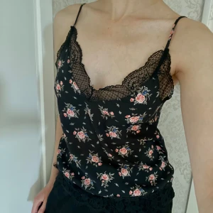 Blommig linnetopp med spets från H&M - Supersöt svart linnetopp från H&M Divided med tunna axelband och blommigt mönster i rosa och beige. Toppen har v-ringning med dekorativ svart spets och en avslappnad passform. Perfekt för att lyfta vilken outfit som helst!