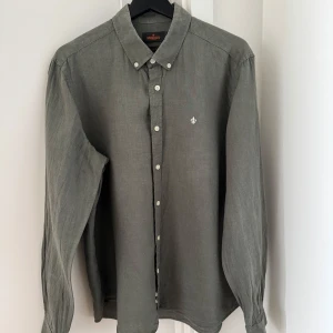 Grön linneskjorta från Morris - Snygg grön linneskjorta från Morris med klassisk krage och knappar framtill. Skjortan har en broderad detalj på bröstet och långa ärmar. Perfekt för en stilren look.