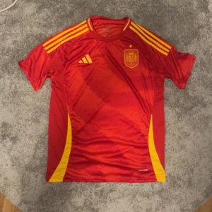 Röd Spansk fotbollströja  - Säljer en röd fotbollströja från Adidas med gula detaljer och korta ärmar. Tröjan har ett emblem på bröstet och tre gula ränder på axlarna. Perfekt för fotbollsfans! ⚽