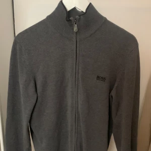 Hugo Boss grå kofta - Grå kofta med dragkedja från Boss Hugo Boss. Koftan har en hög krage och ribbstickade detaljer vid kragen, ärmsluten och nederkanten. En stilren och bekväm kofta som passar perfekt för både vardag och mer formella tillfällen.