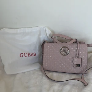 Rosa beige handväska från Guess - Snygg oanvänd handväska från Guess med quiltad design och ett stort metallmärke framtill. Väskan har både handtag och en avtagbar axelrem för flexibilitet. Perfekt för att bära dina dagliga nödvändigheter med stil. Har inte kvitto endast dustbag
