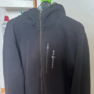 Svart luvtröja från Sail Racing - Snygg svart luvtröja från Sail Racing med dragkedja och huva. Den har en praktisk bröstficka med dragkedja och logotyp på framsidan. Perfekt för kyligare dagar.