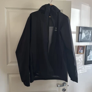 Svart vindjacka från Haglöfs - Säljer en stilren svart vindjacka från Haglöfs. Jackan har en praktisk huva och dragkedja framtill. Perfekt för blåsiga dagar och tillverkad i ett lätt syntetmaterial. Passar bra för utomhusaktiviteter och har en enkel design.
