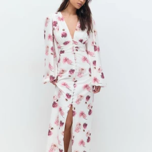 Vit blommig långklänning med v-ringning - Elegant vit långklänning med rosa blommönster och v-ringad design. Klänningen har långa ärmar och knappar framtill som ger en stilren look. Perfekt för sommarens festligheter.