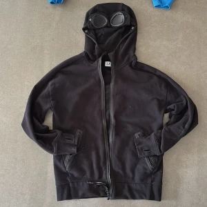 C.P. Hoodie med goggles -  Denna C.P. Company Goggle hoodie i svart är den perfekta kombinationen av stil och komfort. Med sina unika goggle-detaljer på huvan och mjukt material, erbjuder den en avslappnad passform som är idealisk för både vardagsbruk och avslappnade tillfällen. Hoodien har legat i min garderob och nästan aldrig blivit använd, vilket gör den till ett utmärkt tillskott till din 