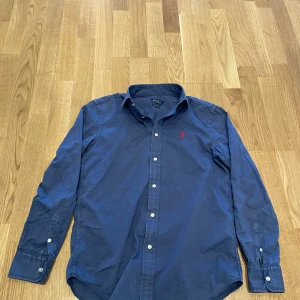 Blå skjorta från Polo Ralph Lauren slim fit - Säljer en klassisk blå skjorta från Polo Ralph Lauren med röd broderad logotyp på bröstet. Skjortan har långa ärmar och knappar framtill. Perfekt för en stilren look.