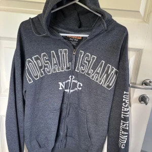 Grå hoodie med dragkedja från Topsail Island - Säljer en grå hoodie med dragkedja och texten 'Topsail Island' tryckt på framsidan och ärmen. Tröjan har en bekväm passform med långa ärmar och en praktisk huva. Perfekt för en avslappnad stil.