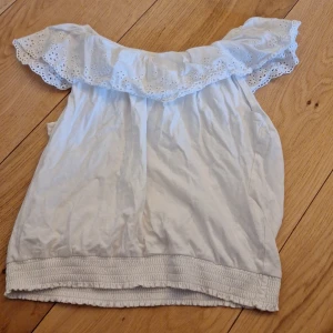 Vit offshoulder topp med spetsdetaljer - Säljer en söt vit offshoulder topp med spetsdetaljer. Toppen har en volangkant och elastisk nederdel för en bekväm passform. Perfekt för varma sommardagar!