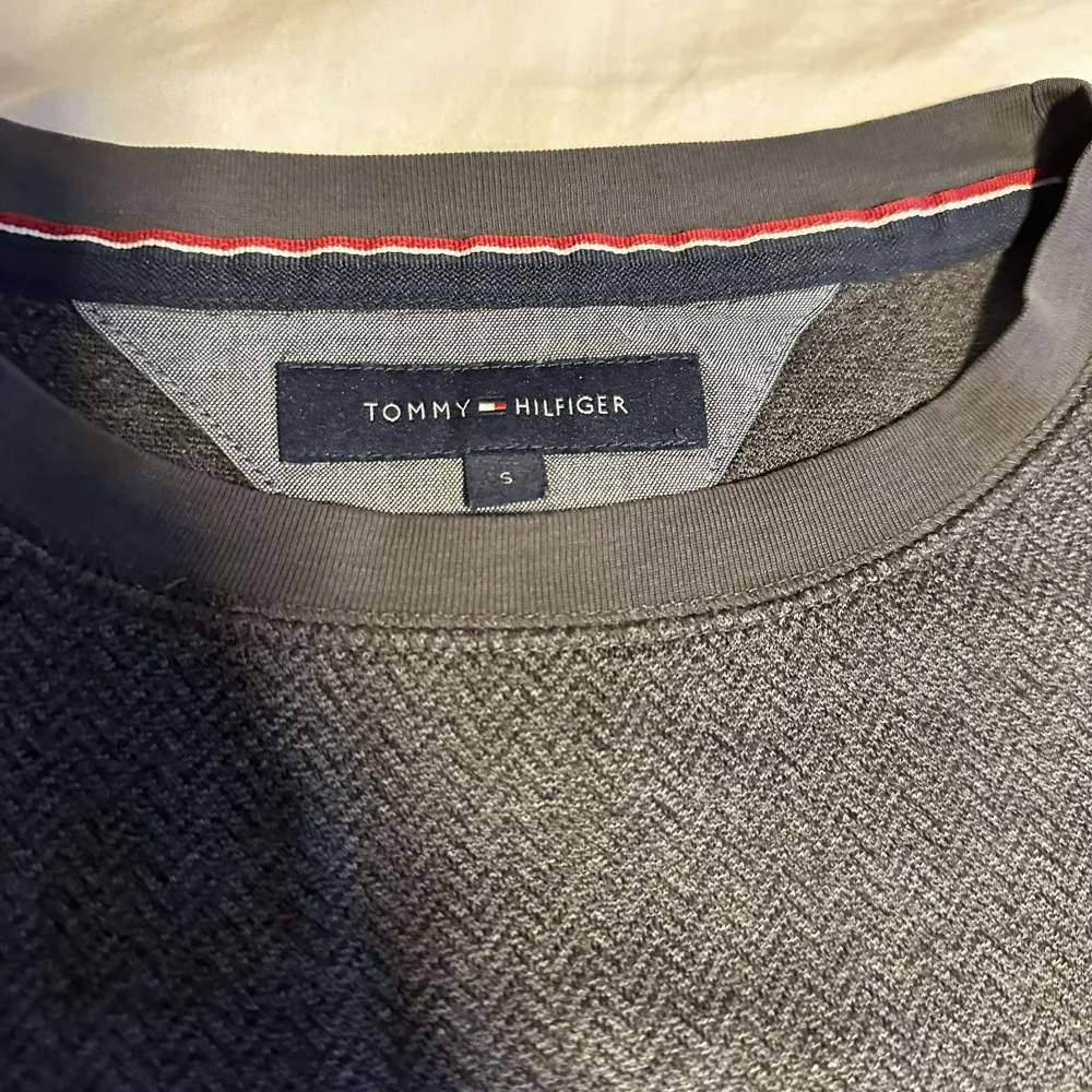 Säljer en stilren grå tröja från Tommy Hilfiger med diskret mönster och rund hals. Tröjan har långa ärmar och en liten logotyp på bröstet. Perfekt för en avslappnad look.. Neuletakit & Villapaidat.