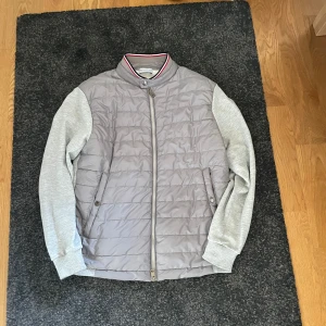 Grå jacka från Moncler - Säljer en stilren grå jacka från Moncler med quiltad framsida och mjuka ärmar. Jackan har en dragkedja framtill och ribbad krage med röd och vit rand. Perfekt för en casual look! Hör av er vid frågor😄