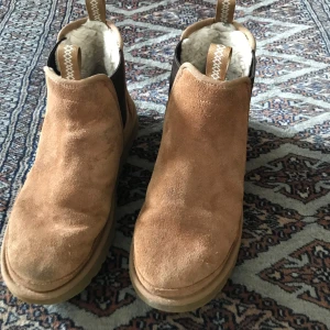Bruna mocka boots från UGG - Säljer ett par bruna mocka boots från UGG med mjukt foder inuti. De har en rund tå och elastiska paneler på sidorna för enkel på- och avtagning. Perfekta till skidbacken eller hockeyträningen. Storlek 44