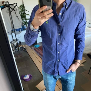 Blå skjorta från Ralph Lauren - Snygg blå skjorta från Ralph Lauren med klassisk krage och knappar framtill. Den har en liten grön broderad logga på bröstet och långa ärmar. Perfekt för en stilren look.