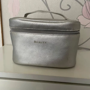 Silverfärgad necessär med handtag - Snygg och praktisk necessär i silverfärgad syntet med ett smidigt handtag och dragkedja. Perfekt för att organisera dina skönhetsprodukter. Ordet 'Beauty' är tryckt på framsidan.