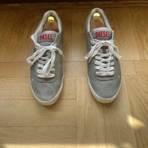 Grå/gröna sneakers från Diesel - Snygga grå/gröna sneakers från Diesel med vit sula och röd under sula. Skorna har en diskret mönstrad yta och röd logga på plösen. Perfekta för en avslappnad stil. Inga defekter.